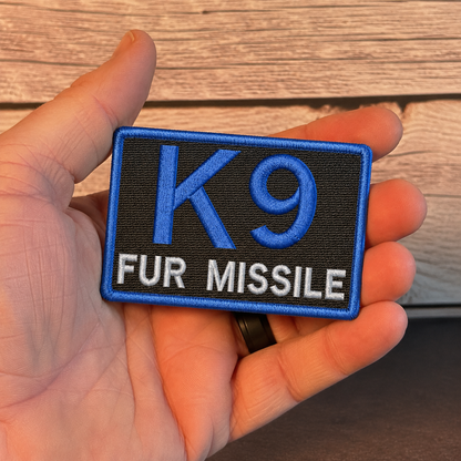 A hand holding a rectangular embroidered patch with a black background, blue border, large blue 'K9' text, and white 'FUR MISSILE' text, against a wood plank background.