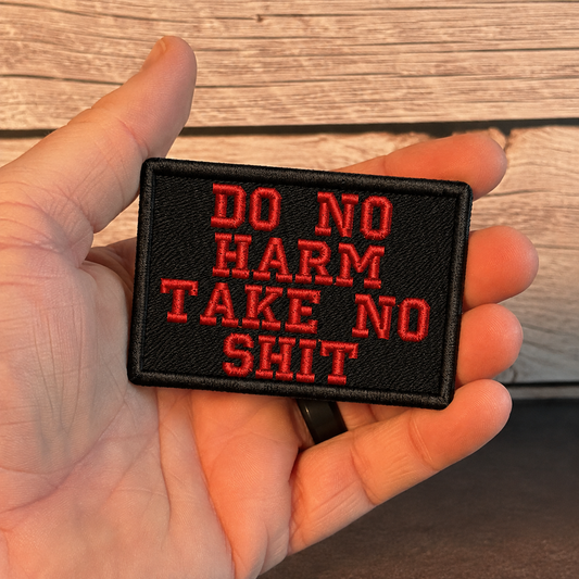 A hand holding a rectangular black embroidered patch with red text that reads 'DO NO HARM TAKE NO SHIT' against a rustic wood plank background.